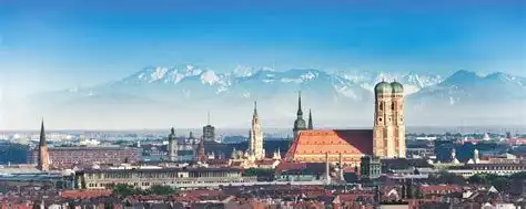 Uno skyline panoramico della città con torri di chiese storiche ed edifici dai tetti rossi, con una catena montuosa innevata sullo sfondo.