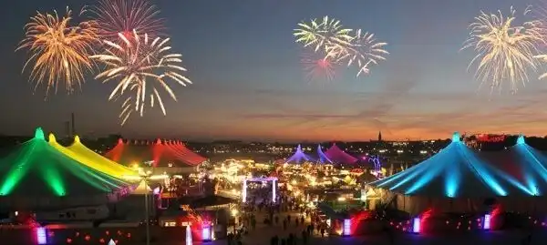 Fuochi d'artificio esplodono su un festival affollato con grandi tende colorate e illuminate al crepuscolo.