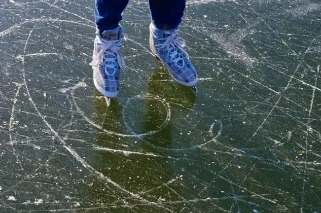 Una persona con pattini da ghiaccio decorati si trova su una superficie di ghiaccio scura coperta da graffi e segni.