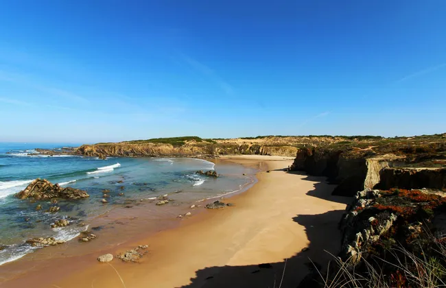 Ampia vista di una spiaggia sabbiosa appartata con onde delicate, circondata da scogliere rocciose sotto un cielo azzurro e limpido.