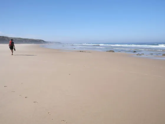 Una persona con un grande zaino cammina a piedi nudi su un'ampia spiaggia sabbiosa, lasciandosi dietro delle impronte.