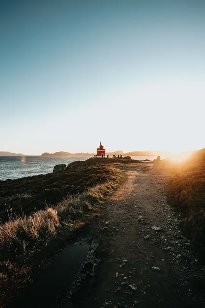 Un gruppo WeRoad si trova accanto a un piccolo faro rosso in cima a una scogliera costiera al tramonto, con un sentiero sterrato in primo piano.