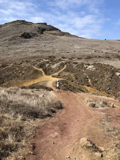 Una persona si trova a un bivio su un sentiero sterrato che si snoda attraverso un paesaggio roccioso e collinare sotto un cielo azzurro con nuvole sparse.