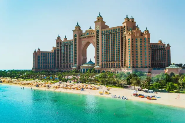 Un grande hotel rosa con un arco centrale si affaccia su una spiaggia di sabbia con palme, ombrelloni gialli e acqua turchese cristallina.