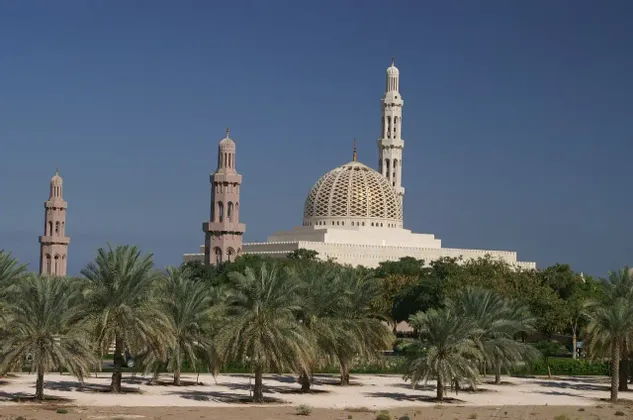 Una grande moschea bianca con una cupola dorata a traliccio e minareti si erge dietro un boschetto di palme sotto un cielo azzurro chiaro.