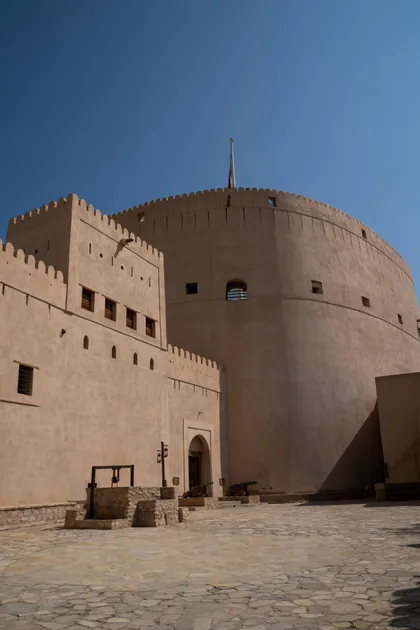 Un imponente forte color sabbia con una torre rotonda domina un cortile in pietra contenente un pozzo e cannoni, sotto un cielo blu limpido.