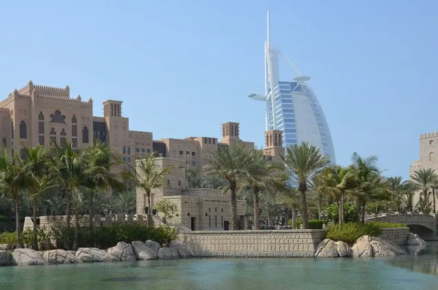 Un grattacielo a forma di vela si erge dietro edifici tradizionali color sabbia e palme, con uno specchio d'acqua calmo in primo piano.
