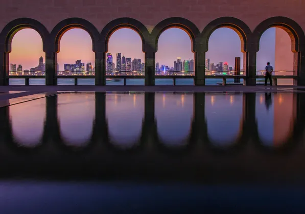Uno scorcio dello skyline cittadino illuminato al crepuscolo è visibile attraverso una serie di archi in pietra, che si riflettono in uno specchio d'acqua.