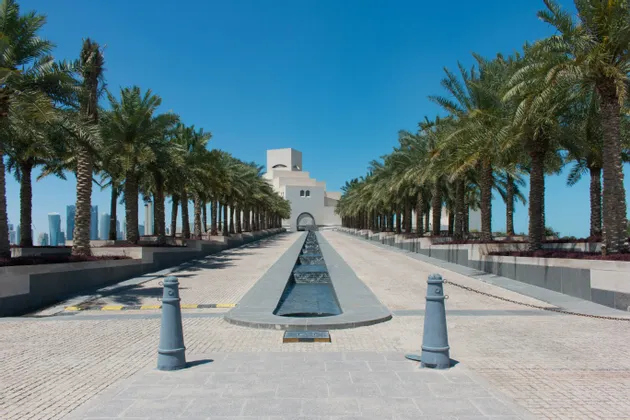 Un'ampia passerella pavimentata, fiancheggiata da palme e con un elemento d'acqua centrale, conduce a un edificio moderno, con lo skyline di una città sullo sfondo.