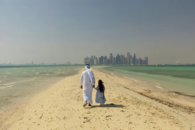 Un uomo in abito tradizionale bianco e una bambina si tengono per mano, camminando su una lingua di sabbia verso il profilo di una città lontana sull'acqua.