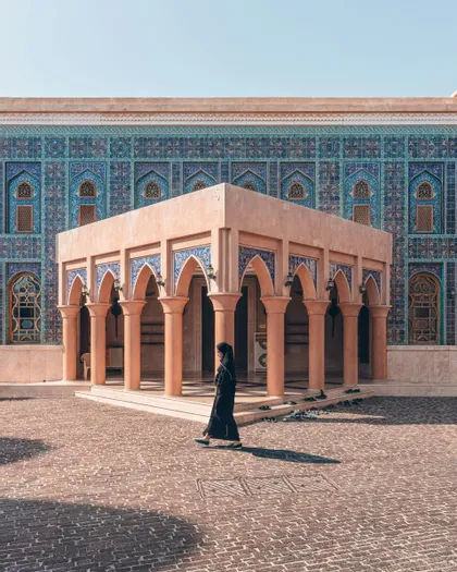 Una persona in abaya nera cammina attraverso un cortile di mattoni davanti a un edificio con piastrelle a mosaico blu e un padiglione ad arco.
