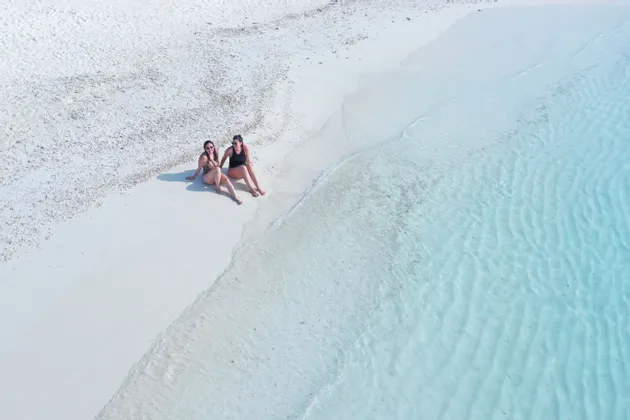 Vista aérea de dos mujeres en traje de baño sentadas en una playa de arena blanca al borde del agua turquesa cristalina.