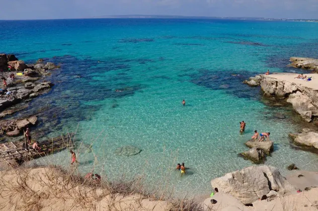 Persone che nuotano e prendono il sole in una caletta rocciosa con acqua limpida e turchese sotto un cielo soleggiato.