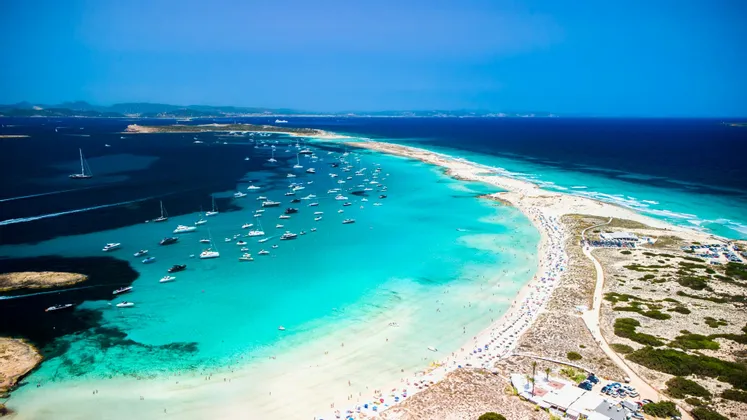 Una vista aerea di una stretta lingua di sabbia con barche ancorate nell'acqua turchese da un lato e il mare blu profondo dall'altro.