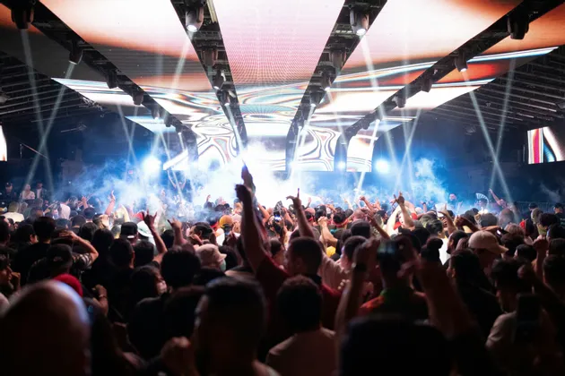 Una grande folla di persone, vista di spalle, che balla con le mani alzate a un concerto con luci brillanti del palco e fumo.