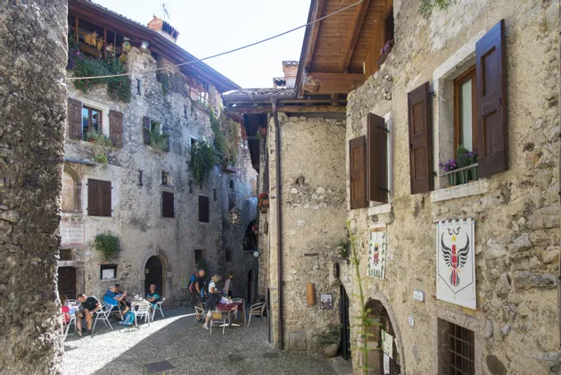Persone sedute ai tavolini all'aperto in un vicolo stretto e acciottolato, illuminato dal sole, tra vecchi edifici in pietra con persiane in legno.