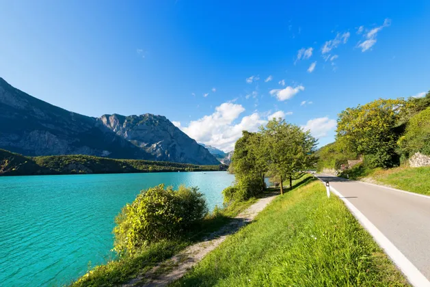 Una strada asfaltata costeggia la riva di un lago turchese brillante, con grandi montagne che si ergono sullo sfondo sotto un cielo azzurro e soleggiato.