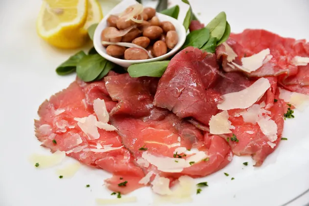 Carpaccio di manzo con scaglie di parmigiano e verdure su un piatto bianco, accanto a una ciotola di fagioli e spicchi di limone fresco.