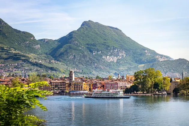Un traghetto naviga su un lago costeggiando una città colorata incastonata ai piedi di grandi montagne verdi sotto un cielo sereno.