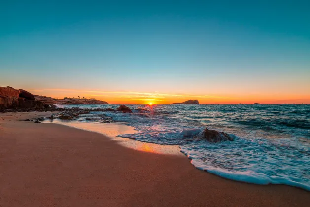 Un tramonto vibrante sull'oceano con onde che lambiscono una spiaggia sabbiosa accanto a una costa rocciosa.