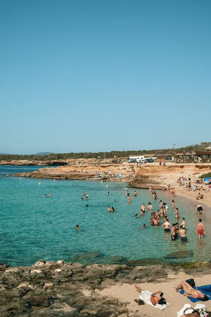 Una folla di persone che nuota e si rilassa su una spiaggia rocciosa e assolata con acqua limpida e turchese.