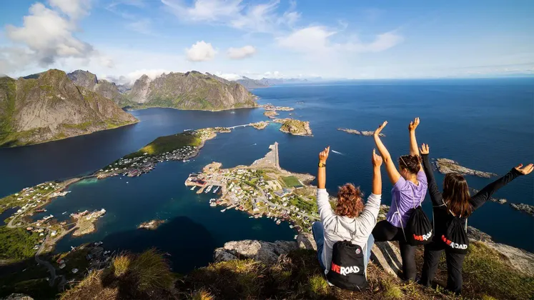 Eine WeRoad-Gruppe dreier Frauen sitzt mit dem Rücken zur Kamera auf einer Klippe und blickt auf eine Küstenstadt, Berge und einen Fjord.