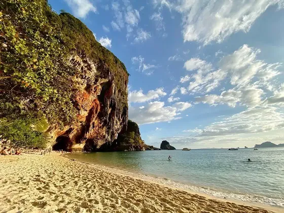 Eine große Felsklippe mit einer Höhle grenzt an einen Sandstrand, an dem Menschen unter einem teilweise bewölkten Himmel schwimmen und entspannen.