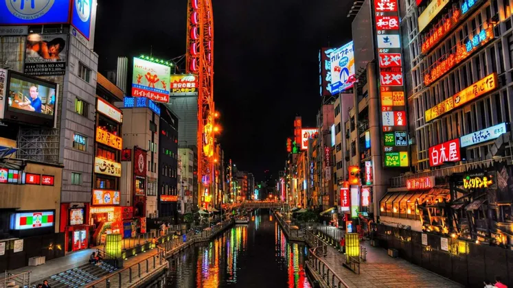 Un canale cittadino di notte fiancheggiato da edifici con insegne al neon giapponesi colorate che si riflettono sull'acqua.