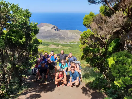 Eine WeRoad-Gruppenreise posiert auf einem von Bäumen gesäumten Feldweg mit Blick auf eine Vulkanlandschaft und das Meer.