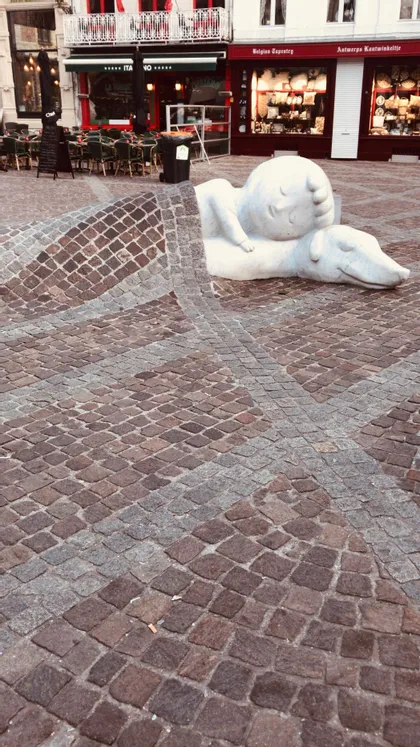 Una scultura bianca di un bambino che dorme su un cane, con un selciato di ciottoli che funge da coperta su di loro in una piazza della città.