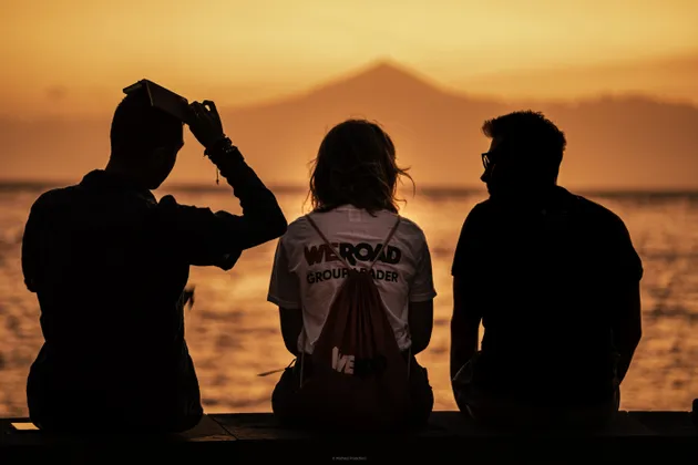 Trois personnes d'un voyage en groupe WeRoad sont assises en silhouette sur une jetée, contemplant un coucher de soleil orangé sur l'eau avec une montagne au loin.