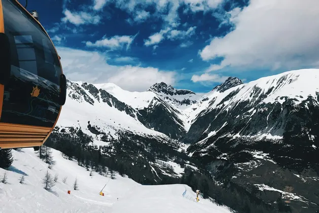 Vista da una funivia gialla su una vasta catena montuosa innevata sotto un cielo blu nuvoloso.