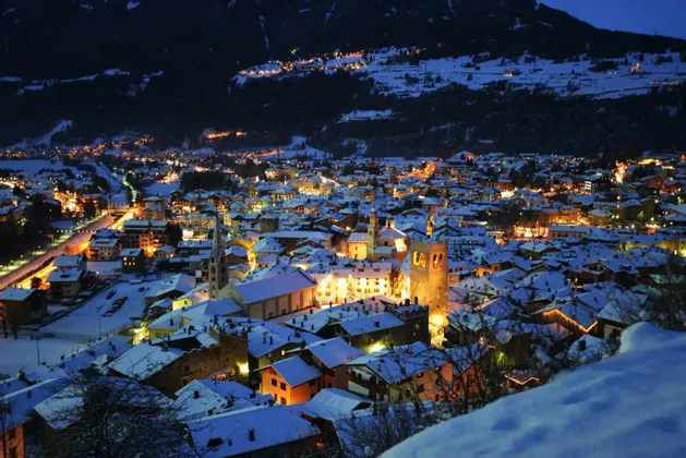 Una città alpina con tetti innevati è illuminata a giorno di notte, incastonata ai piedi di una montagna scura.