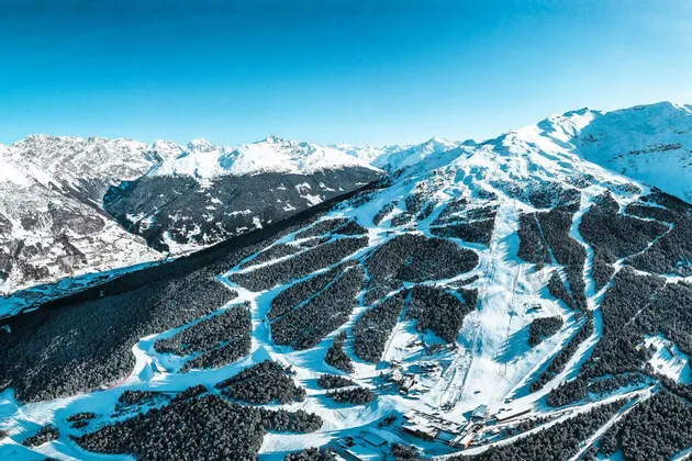 Una vista aerea di una stazione sciistica con diverse piste che attraversano una foresta montana innevata sotto un cielo sereno.