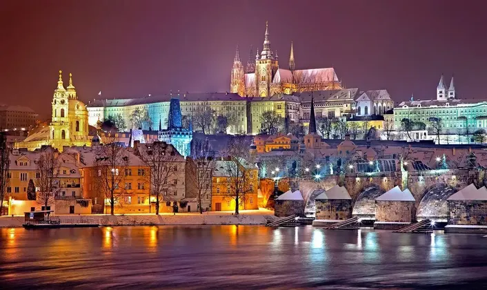 Une ville historique enneigée avec un château et un pont illuminés la nuit, les lumières se reflétant sur la rivière en contrebas.