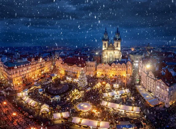 Vue aérienne d'un marché de Noël animé sur une place de ville la nuit, avec de la neige tombant sur des stands illuminés et un grand sapin.