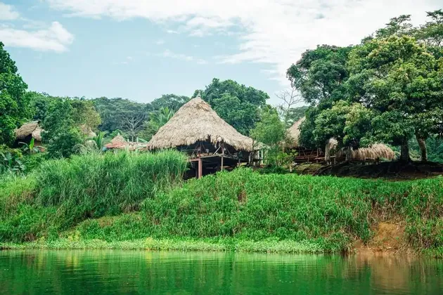 Un villaggio di capanne con tetti di paglia si trova su una rigogliosa sponda verde di fiume, circondato da densi alberi tropicali.