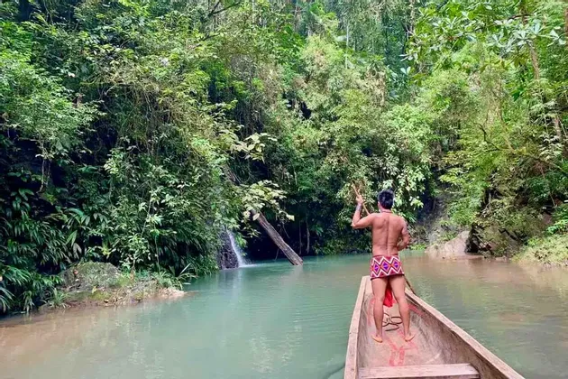 Un uomo in pantaloncini tradizionali si trova a prua di una canoa di legno, navigando un fiume tranquillo attraverso una giungla verde e fitta.