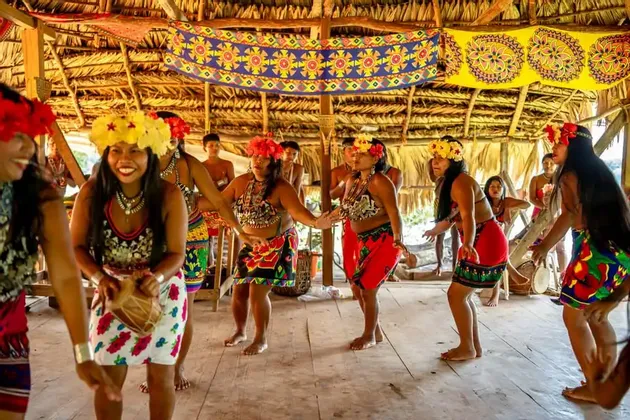 Un gruppo di donne indigene che indossano abiti tradizionali colorati e corone di fiori balla all'interno di una capanna con il tetto di paglia.