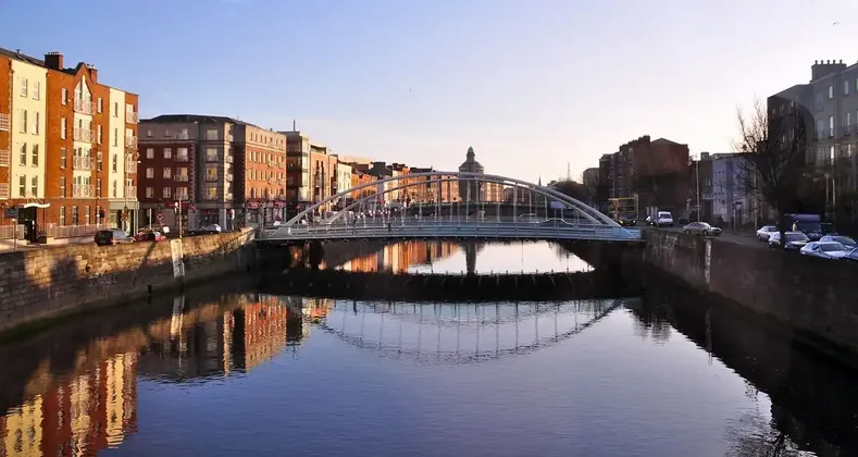 Un moderno ponte ad arco attraversa un fiume calmo in una città all'alba, il suo riflesso che specchia gli edifici lungo il lungofiume.