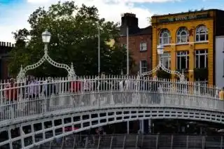 Gente attraversa un affollato e ornato ponte pedonale bianco, con un edificio giallo e alberi verdi sullo sfondo, sotto un cielo azzurro.