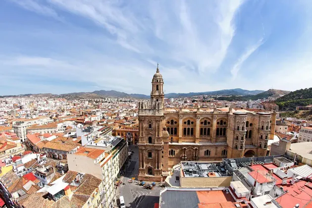 Veduta aerea di una grande cattedrale con campanile circondata da un paesaggio urbano di tetti in terracotta e montagne lontane.
