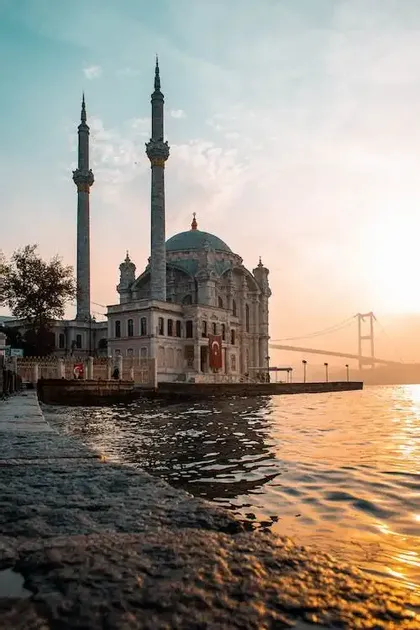 Una mezquita ornamentada con dos minaretes se alza junto al agua al atardecer, con un puente a lo lejos y reflejos en el agua.