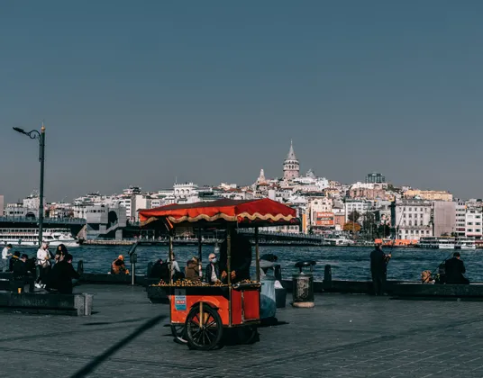 Un carrito rojo de comida callejera en un paseo marítimo con un paisaje urbano y una torre visibles al fondo al otro lado del agua.