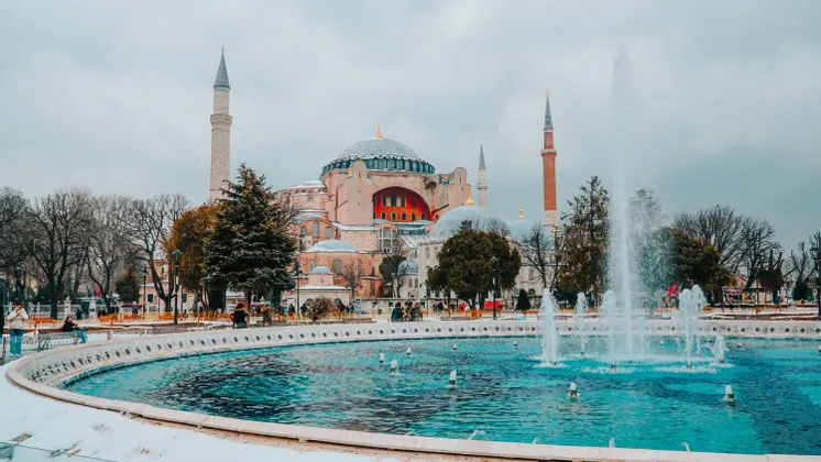 Una gran mezquita con cúpulas y minaretes se alza detrás de una fuente circular con agua turquesa en un día nevado.