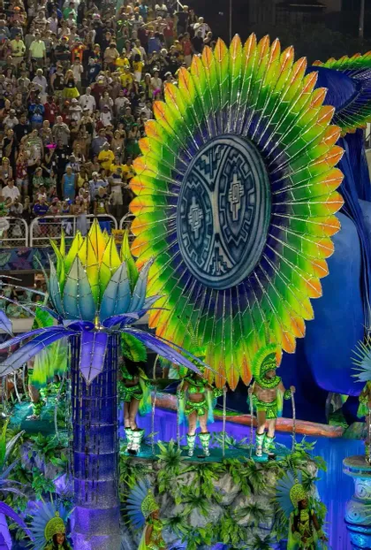 Artisti con elaborati costumi verdi su un grande carro allegorico colorato e decorato con piume, durante una parata notturna osservata dalla folla.