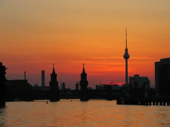 Un horizon urbain avec un pont et une haute tour se découpant sur un coucher de soleil orangé au-dessus d'un fleuve.