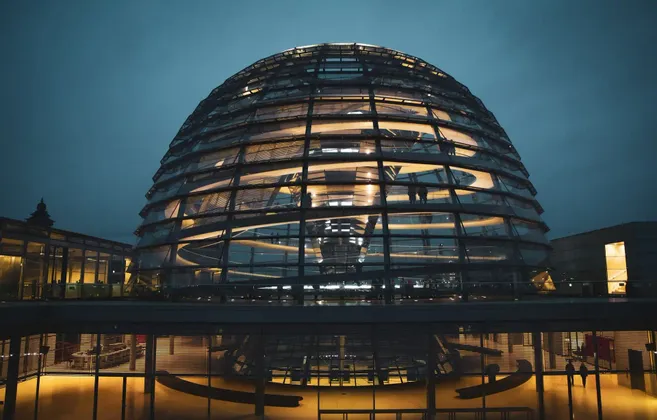 Le dôme de verre illuminé d'un bâtiment moderne la nuit, avec des silhouettes de personnes visibles sur une passerelle intérieure.