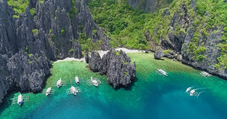 Eine Luftaufnahme von Auslegerbooten, die auf türkisfarbenem Wasser in einer abgelegenen Bucht mit zerklüfteten Kalksteinfelsen und einem weißen Sandstrand schwimmen.
