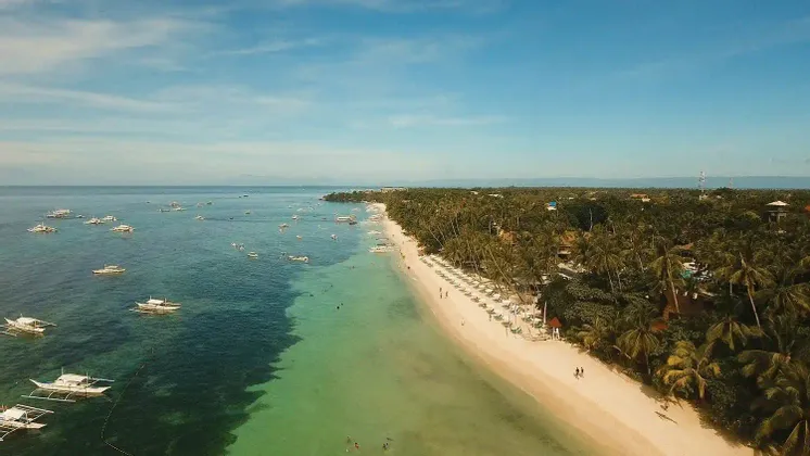 Luftaufnahme einer tropischen Küste mit weißem Sandstrand, dichten Palmen und zahlreichen weißen Auslegerbooten auf dem Wasser.
